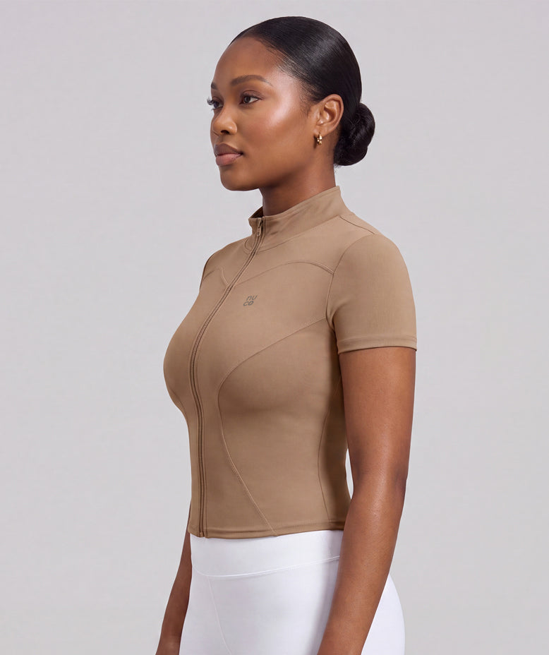 A woman in profile wearing a fitted short-sleeve athletic top in tan, paired with white leggings, against a plain light gray studio background.