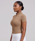 A woman in profile wearing a fitted short-sleeve athletic top in tan, paired with white leggings, against a plain light gray studio background.