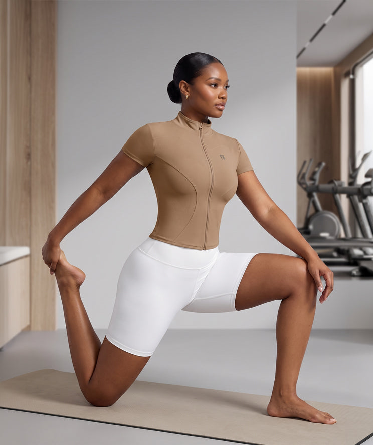 A woman in a gym wearing a coffee toffee brown short-sleeve athletic top and white fitted shorts, performing a lunge stretch on a yoga mat with exercise equipment in the background.